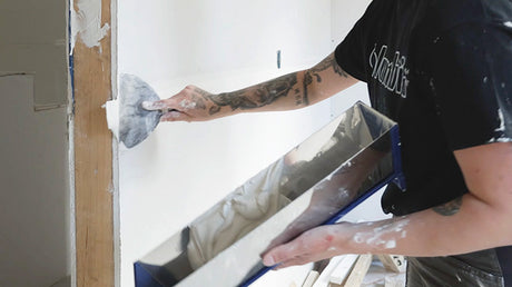 Drywaller applying joint compound over drywall tape with a joint knife while holding a mud pan